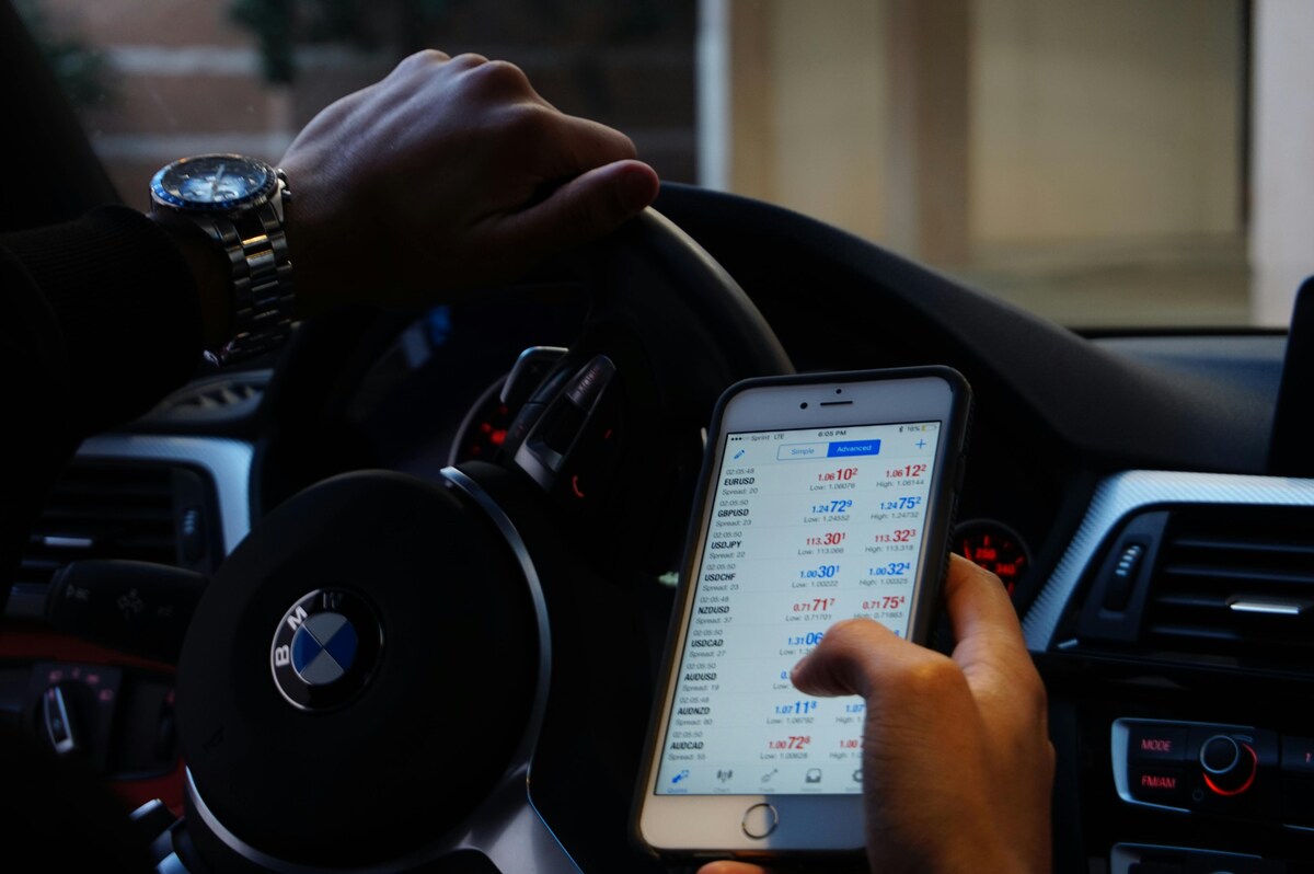 A person checking forex trading rates on a smartphone while driving a BMW.